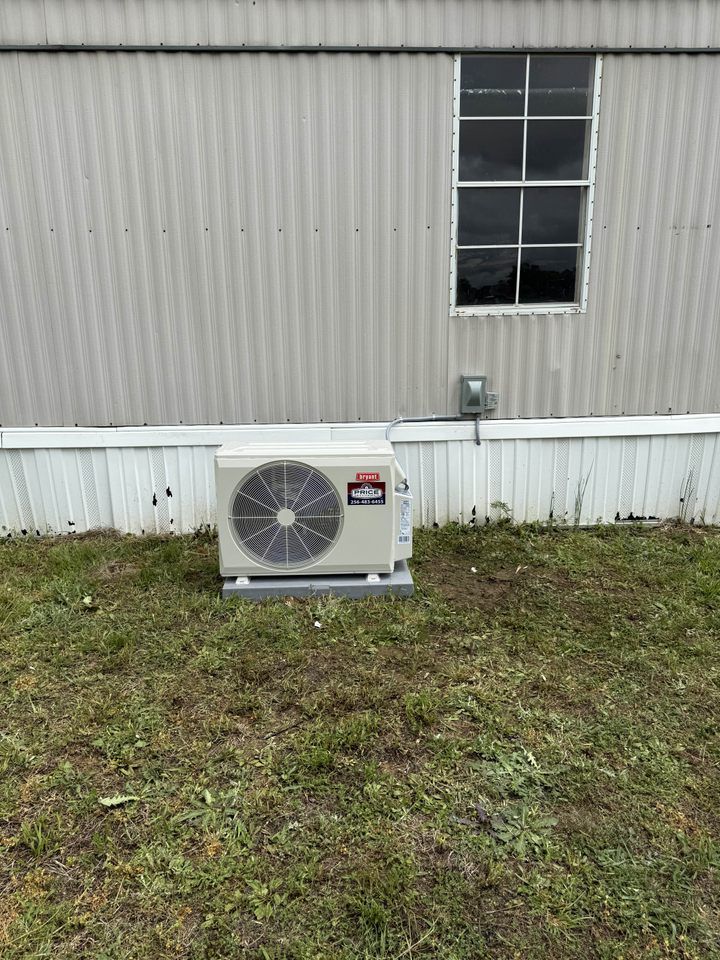 An outdoor mini-split air conditioning unit is installed on a concrete pad next to the skirting of a light-colored mobile home, with a window visible above the unit.