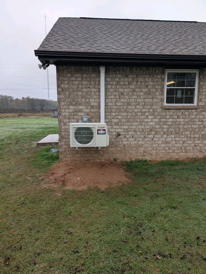An outdoor ductless mini-split heat pump unit is mounted on the side of a brick building. The unit is white and has a Mitsubishi logo. A white conduit runs vertically up the wall from the unit. The ground below the unit is bare dirt, surrounded by grass. The background shows a field and trees under a cloudy sky.