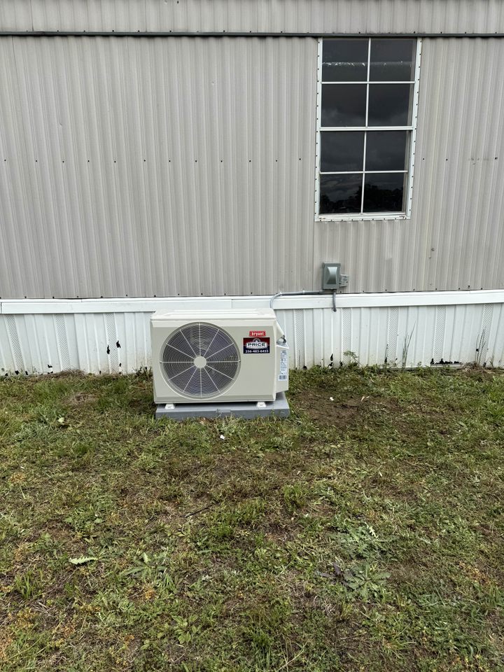 An outdoor mini-split air conditioning unit is installed on a concrete pad next to the skirting of a light-colored mobile home, with a window visible above the unit.